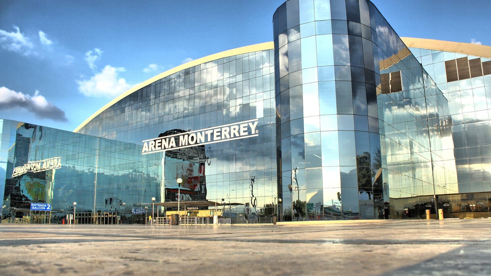 Fachada de vidrio de la Arena Monterrey con su letrero y su arco blanco, vista desde la explanada.